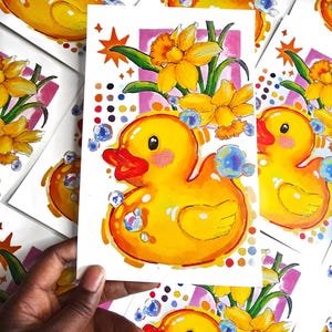 Puede incluir: Ilustraci&oacute;n vibrante de un patito de goma amarillo con pico rojo y burbujas. Narcisos y un marco rosa enmarcan el pato. La obra de arte est&aacute; en una tarjeta blanca, con otras tarjetas similares en el fondo.