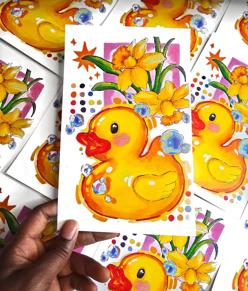 Puede incluir: Ilustraci&oacute;n vibrante de un patito de goma amarillo con pico rojo y burbujas. Narcisos y un marco rosa enmarcan el pato. La obra de arte est&aacute; en una tarjeta blanca, con otras tarjetas similares en el fondo.