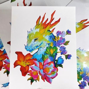 May include: A colorful art print featuring a dragon's head with a vibrant color palette. The dragon is surrounded by flowers in shades of orange, pink, and purple. The artwork is on white paper.