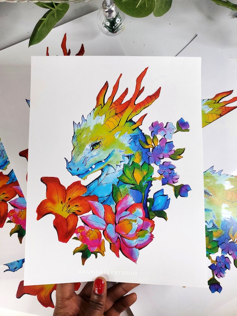 May include: A colorful art print featuring a dragon's head with a vibrant color palette. The dragon is surrounded by flowers in shades of orange, pink, and purple. The artwork is on white paper.