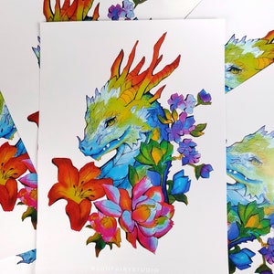 May include: A colorful art print featuring a dragon's head with a vibrant color palette. The dragon is surrounded by flowers in shades of red, pink, purple, and green. The text "@SUNFAIRYSTUDIO" is printed at the bottom.