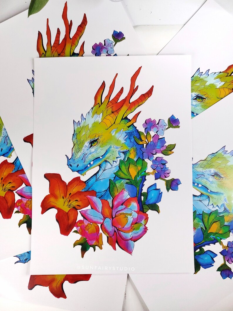 May include: A colorful art print featuring a dragon's head with a vibrant color palette. The dragon is surrounded by flowers in shades of red, pink, purple, and green. The text "@SUNFAIRYSTUDIO" is printed at the bottom.