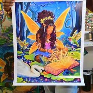 May include: A vibrant fantasy art print featuring a fairy with orange wings, sitting on a lily pad. The fairy is surrounded by hummingbirds, a swan, and a frog. An open book glows with light. The artwork is set against a blue and green background.