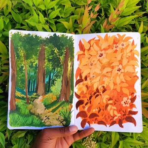 May include: A sketchbook open to two landscape paintings. One depicts a forest scene with a path, the other a cluster of orange and brown leaves. The paintings are held against a backdrop of green foliage.