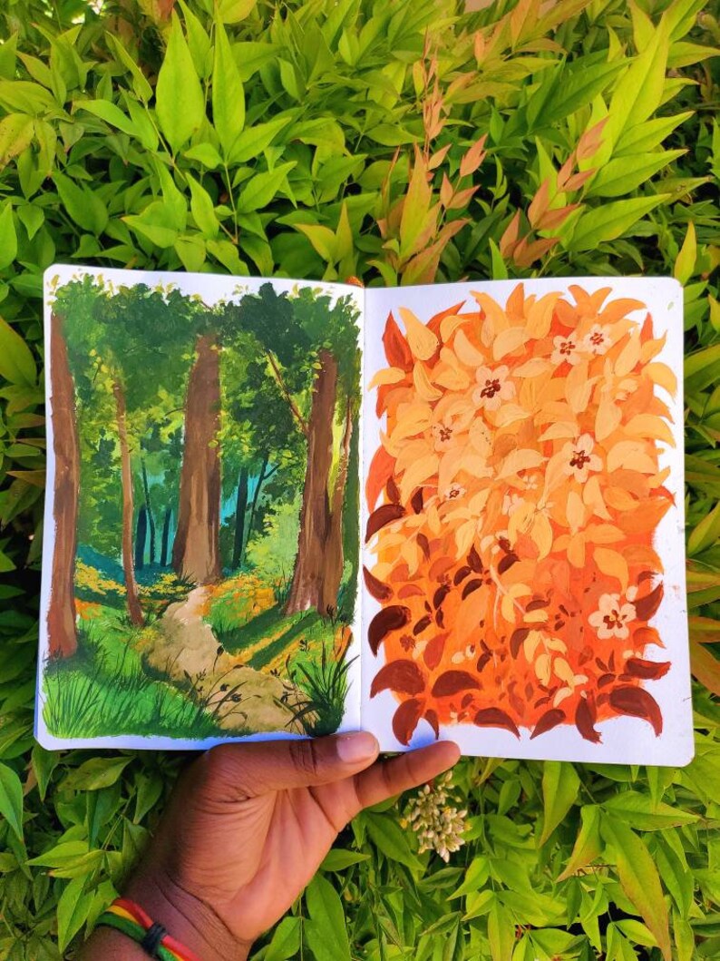May include: A sketchbook open to two landscape paintings. One depicts a forest scene with a path, the other a cluster of orange and brown leaves. The paintings are held against a backdrop of green foliage.