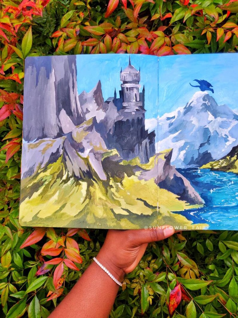 Puede incluir: Una pintura de paisaje que presenta un castillo en una monta&ntilde;a rocosa, un pico nevado y un drag&oacute;n azul. La obra de arte est&aacute; ambientada contra un cielo azul y una masa de agua. La pintura se sostiene contra un fondo de follaje verde y rojo.