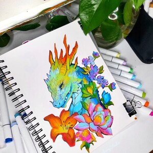 May include: A colorful dragon illustration in a spiral-bound sketchbook. The dragon is depicted with blue, green, and orange scales, surrounded by vibrant flowers in shades of orange, pink, and purple. The artist's signature is visible.
