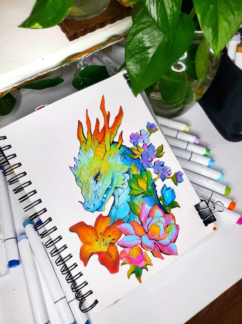 May include: A colorful dragon illustration in a spiral-bound sketchbook. The dragon is depicted with blue, green, and orange scales, surrounded by vibrant flowers in shades of orange, pink, and purple. The artist's signature is visible.