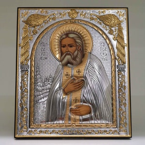 Może przedstawiać: Ikona srebrna i złota przedstawiająca brodatego mężczyznę w białej szacie na złotym tle. Ikona jest oprawiona w czarną drewnianą ramę z złotymi i srebrnymi akcentami. Tekst na ikonie brzmi "ПРП СЕ Р ФМ" i "СВ РОВ СК УЧ".