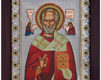 May include: A religious icon depicting Saint Nicholas, a traditional Christian figure, in a red and green robe holding a book. The icon is framed in a silver and gold ornate frame with floral designs.