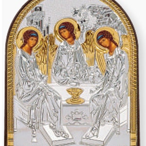 May include: A silver religious icon depicting three figures with halos, seated around a table. The figures are in detailed silver robes, with gold accents and a dark brown frame. The icon has an arched top and intricate detailing.
