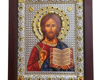 May include: A framed religious icon depicting Jesus Christ holding a book with Cyrillic text. The icon is set against a silver background with gold accents and intricate floral designs.