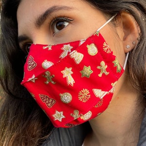 Puede incluir: Una mujer con una mascarilla roja con un patrón navideño. La mascarilla tiene estrellas doradas, árboles, adornos y hombres de jengibre.