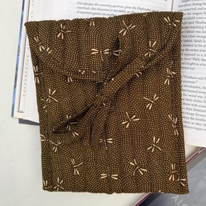 May include: A brown fabric book sleeve with a dragonfly pattern and a knotted closure. The sleeve is lying on top of an open book and a tablet.
