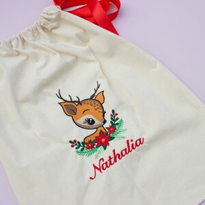 May include: A white canvas gift bag with a red ribbon. The bag has an embroidered design of a cartoon deer with a red bow and green holly. The name "Nathalia" is embroidered in red below the deer.