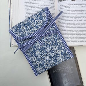 May include: A blue floral print Kindle e-reader case with a gingham trim, lying open on a book. The e-reader displays the title "ALCHEMISED". The case is designed to protect and carry e-readers.