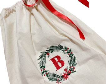 May include: A white canvas gift bag with a red ribbon and a green and red embroidered wreath with the letter "B" in the center.