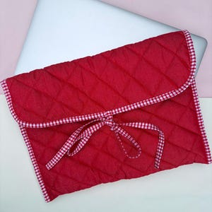 May include: Red quilted laptop sleeve with a white and red gingham trim and matching tie closure. The sleeve is designed to hold a laptop and is shown with a silver laptop inside. The background is a light pink and white.