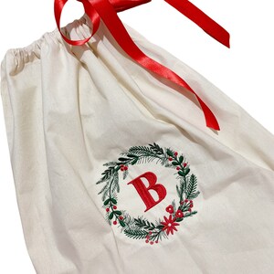 May include: A white canvas gift bag with a red ribbon and a green and red embroidered wreath with the letter "B" in the center.
