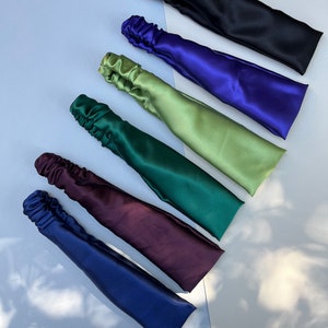 May include: Six satin headbands in different colours: black, purple, green, dark green, burgundy, and navy blue. The headbands are arranged in a row, with the black headband at the top and the navy blue headband at the bottom.