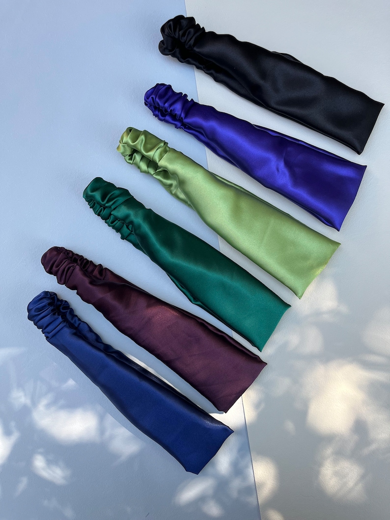May include: Six satin headbands in different colours: black, purple, green, dark green, burgundy, and navy blue. The headbands are arranged in a row, with the black headband at the top and the navy blue headband at the bottom.