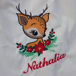 May include: A white canvas tote bag with an embroidered image of a brown deer with large eyes and red flowers. The name "Nathalia" is embroidered in red below the deer.