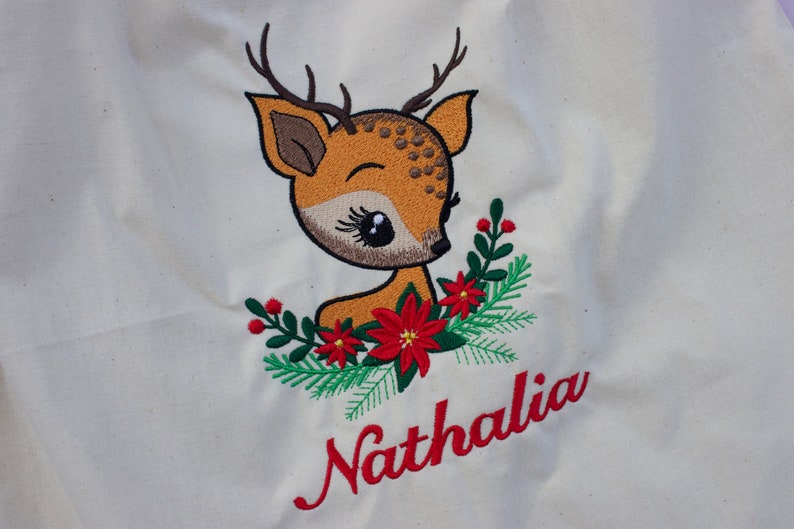 May include: A white canvas tote bag with an embroidered image of a brown deer with large eyes and red flowers. The name "Nathalia" is embroidered in red below the deer.