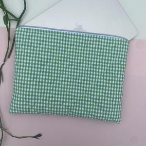 May include: A green and white gingham laptop sleeve with a silver zipper. The sleeve is made of a quilted fabric and is partially covering a silver laptop. The background is a mix of pink and white.