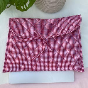 May include: Red and white gingham laptop sleeve with a quilted design. The sleeve has a flap closure and a matching tie. The laptop sleeve is designed to protect a laptop.
