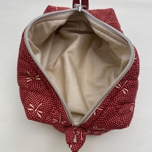 May include: Red zippered pouch with a white dragonfly pattern and a beige lining. The pouch is open and the zipper is pulled down.