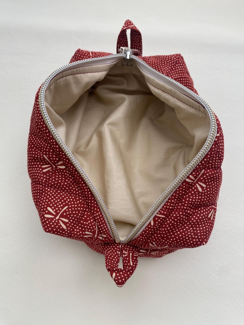 May include: Red zippered pouch with a white dragonfly pattern and a beige lining. The pouch is open and the zipper is pulled down.