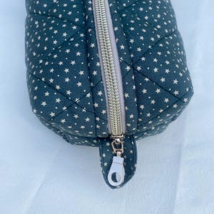 Può includere: Un beauty case trapuntato in tessuto verde scuro, decorato con piccole stelle bianche. La borsa presenta una chiusura con cerniera argentata e un tirante abbinato. Le cuciture creano un motivo a rombi sulla superficie della borsa.