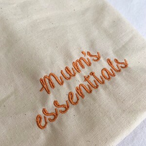 Puede incluir: Una bolsa de lona beige con el texto "Mummy's essentials" bordado en hilo naranja. El texto est&aacute; en una fuente cursiva.