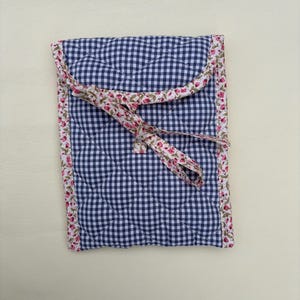 May include: A quilted fabric case with a blue and white gingham pattern. The edges are trimmed with a floral print, and a matching floral ribbon ties the case closed. The case has a diamond-shaped quilted pattern.