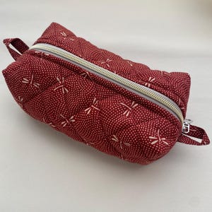 May include: A red quilted zippered pouch with a white dragonfly pattern. The pouch has a gold zipper and a small loop handle.