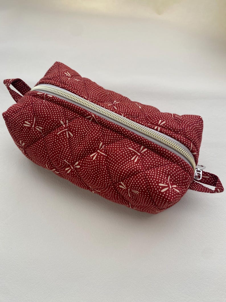 May include: A red quilted zippered pouch with a white dragonfly pattern. The pouch has a gold zipper and a small loop handle.