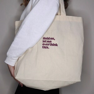 Puede incluir: Una bolsa de lona blanca con el texto "Hold on, let me overthink this." impreso en granate.