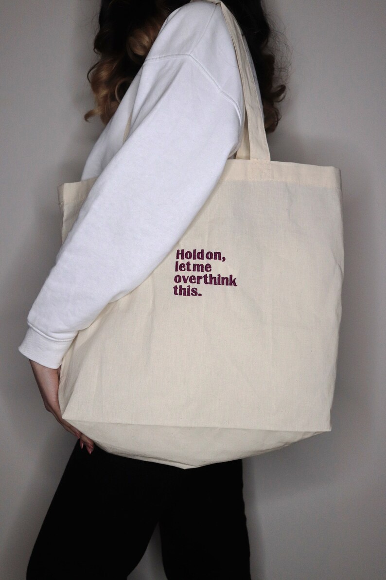 Puede incluir: Una bolsa de lona blanca con el texto "Hold on, let me overthink this." impreso en granate.