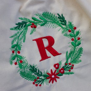 May include: A white fabric bag with a red embroidered wreath featuring green leaves, red berries, and a red letter "R" in the center.
