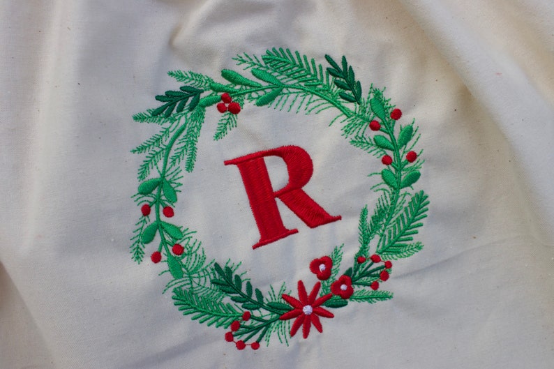 May include: A white fabric bag with a red embroidered wreath featuring green leaves, red berries, and a red letter "R" in the center.