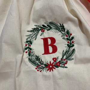 May include: A white canvas Christmas gift bag with a red ribbon. The bag is embroidered with a green wreath with red berries and a red letter "B" in the center.