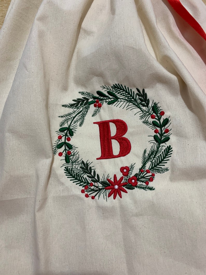 May include: A white canvas Christmas gift bag with a red ribbon. The bag is embroidered with a green wreath with red berries and a red letter "B" in the center.