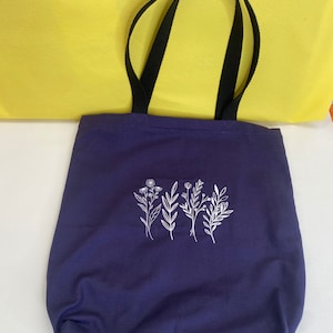 Puede incluir: Una bolsa de tela de color morado oscuro con asas negras y un diseño floral blanco.