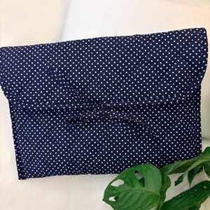 May include: A navy blue clutch with a white polka dot pattern. The clutch is rectangular with a folded flap closure. The fabric appears to be soft and slightly textured. A green plant with large leaves is visible in the corner.