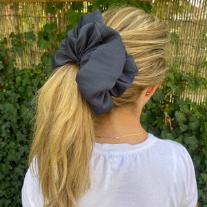 Puede incluir: Una scrunchie de satén gris oscuro asegura una cola de caballo rubia. El accesorio para el cabello de gran tamaño está hecho de una tela suave y fluida. El fondo presenta follaje verde y una valla de madera.