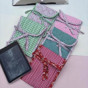 May include: A collection of fabric e-reader cases in various colors and patterns, including green, pink, and red gingham, and floral prints. Each case has a coordinating floral trim and a ribbon closure. An e-reader with the text "ALCHEMISED" is visible.