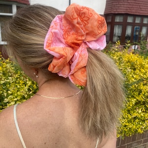 May include: A pink and orange floral patterned hair scrunchie. The scrunchie is tied around a ponytail.