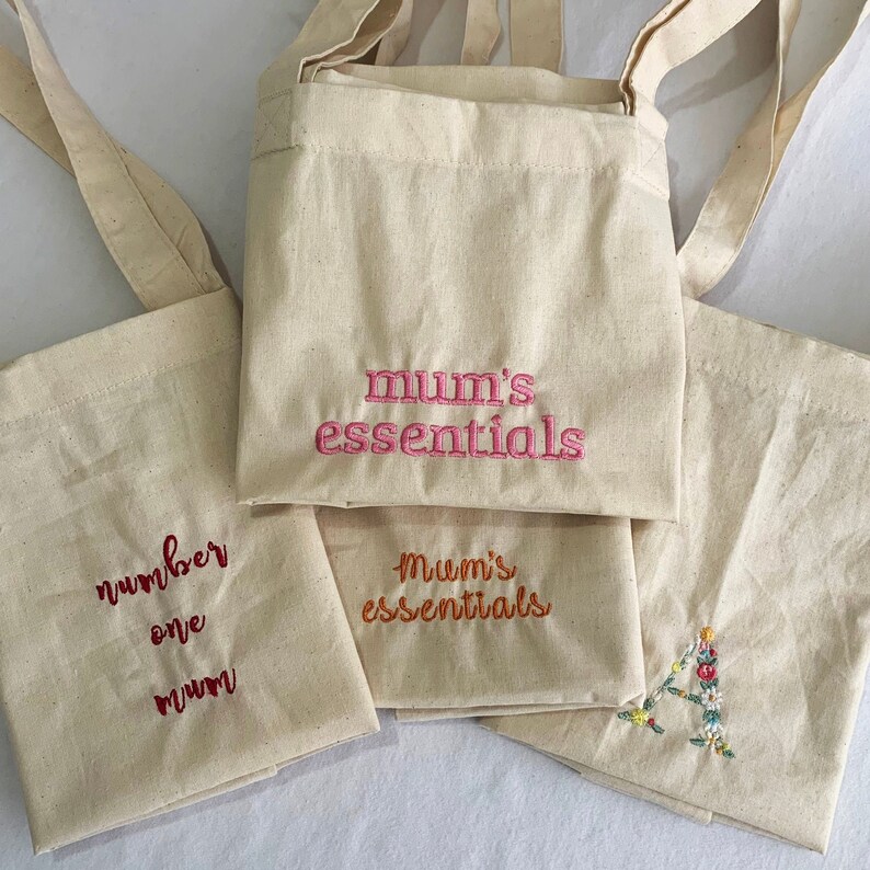 Puede incluir: Cuatro bolsas de lona beige con diferentes dise&ntilde;os bordados. La bolsa de arriba tiene "mum's essentials" bordado en rosa. La bolsa de abajo tiene "mum's essentials" bordado en naranja. La bolsa de la izquierda tiene "number one mum" bordado en rojo. La bolsa de la derecha tiene un dise&ntilde;o floral bordado en naranja.