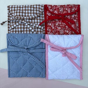 May include: Four quilted fabric pouches in a square arrangement. The pouches feature different patterns: brown and white gingham, red floral, blue and white gingham, and white eyelet. Each pouch has a flap closure and a matching tie.