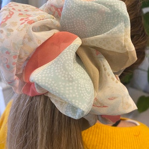 May include: A large, colourful hair scrunchie made of soft, sheer fabric with a floral pattern. The scrunchie is tied around a ponytail and features shades of pink, beige, and light blue.